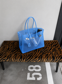 Sac Jelly Firkin Croco Azure bleu posé sur un banc à motif tigre, décor urbain avec marquage au sol